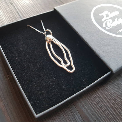 Pictured is a sterling silver vulva necklace pictured resting inside it's box over a charred wooden board.
