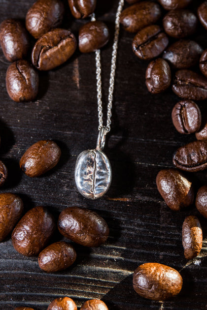 Resting on a charred wooden board sits a sterling silver coffee bean necklace surrounded in roasted coffee beans
