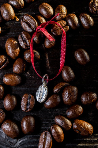A sterling silver coffee bean charm pendant upon a red ribbon loop is resting on a wooden board that is filled with roasted coffee beans.