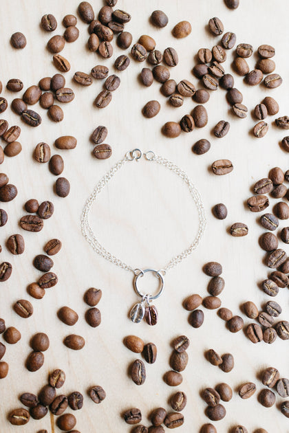Laying flat on a table adorned by coffee beans sits a coffee bean bracelet with a silver ring holding two coffee beans