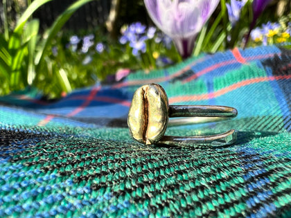 A handmade sterling silver adjustable ring with a polished brass coffee bean is resting on a Scottish tartan kilt