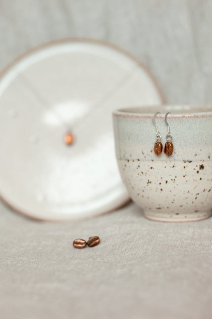 a set of red bronze coffee bean drop earrings resting on the lip of a coffee cup, with red bronze coffee bean stud earrings resting on a linen cloth below and a red bronze coffee bean necklace sitting upon a plate in the background of the image.