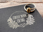 sitting upon a black jewellery box sits a sterling silver coffee bean ring with a polished brass coffee bean