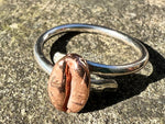 sitting on a stone slate sits a sterling silver ring with a red bronze coffee bean on it