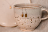 resting from a cup sits a pair of brass coffee bean earrings with silver hooks from a wide angle