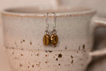 resting from a cup sits a pair of brass coffee bean earrings with silver hooks